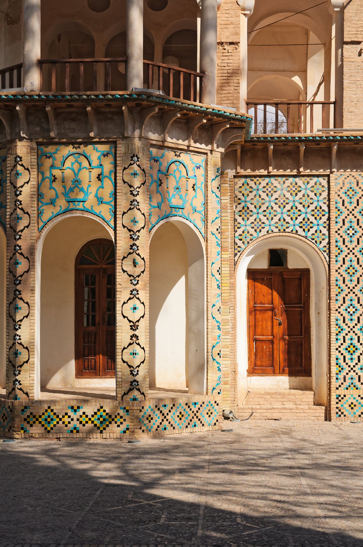 Tiled arcades at the Shazdeh Garden in Mahan, near Kerman: brick piers framed by turquoise floral tilework and a band of geometric mosaic, with arched recesses and wooden doors in shadow