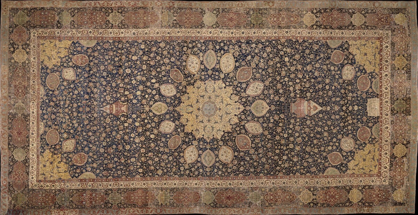 The Ardabil carpet: a golden sunburst medallion with sixteen pendants and two mosque lamps on an indigo field of flowering vines, within a complete border system, an inscription cartouche at one end.