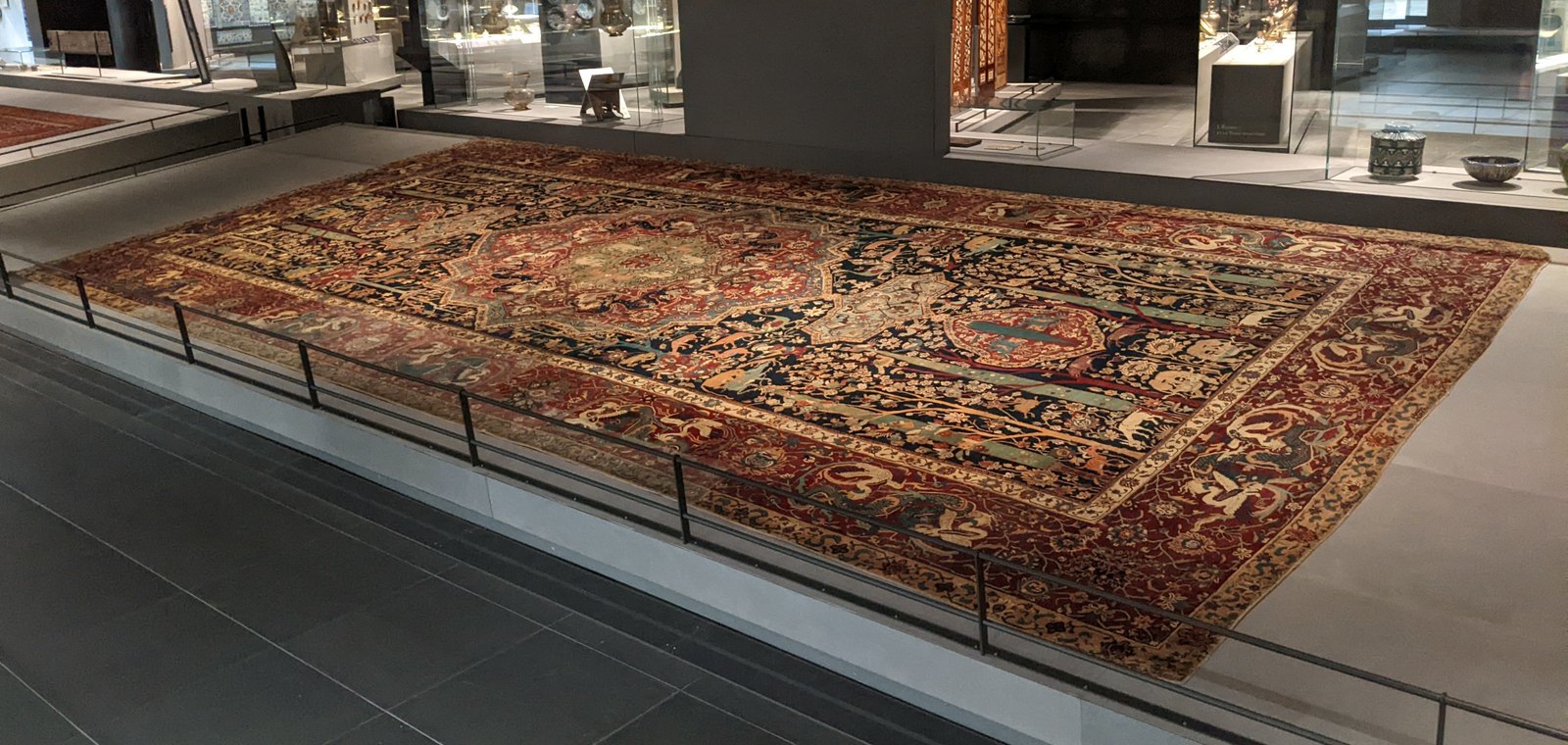 The Mantes carpet lying behind a rail in the Louvre&rsquo;s Islamic art galleries, display cases glowing behind it.