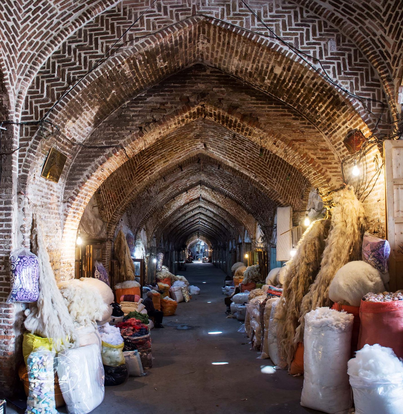 A vaulted brick row of the Tabriz Grand Bazaar receding in perspective, lined with sacks of raw and dyed wool, the herringbone brickwork of the arched ceiling lit by hanging bulbs