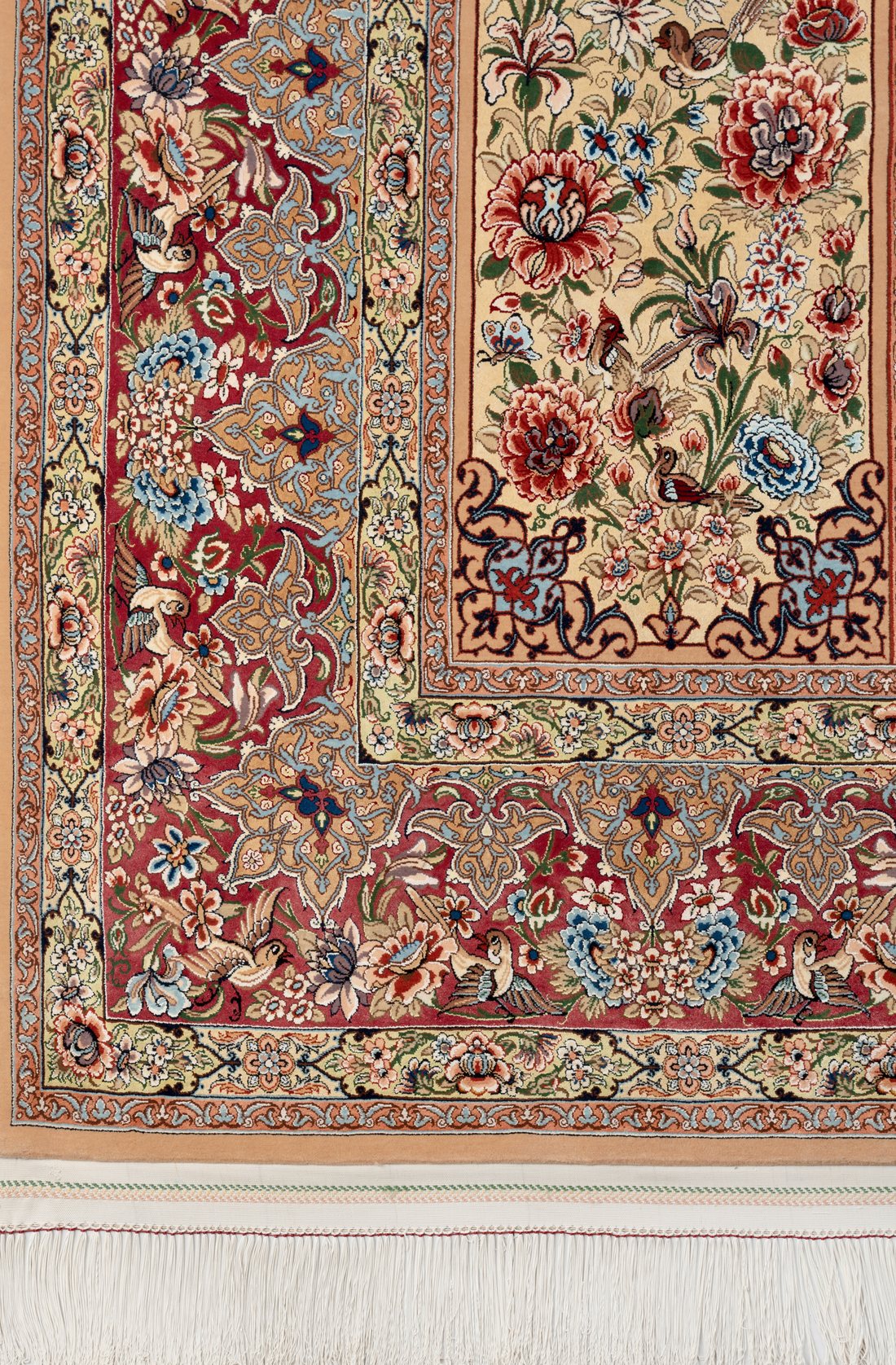 Corner detail showing the border and the Gol o Bolbol panel: the red main band scrolling with palmettes and small birds, the pistachio-green minor band and guard stripes, beside the cream flower-and-bird panel with its large blossoms and iris forms.