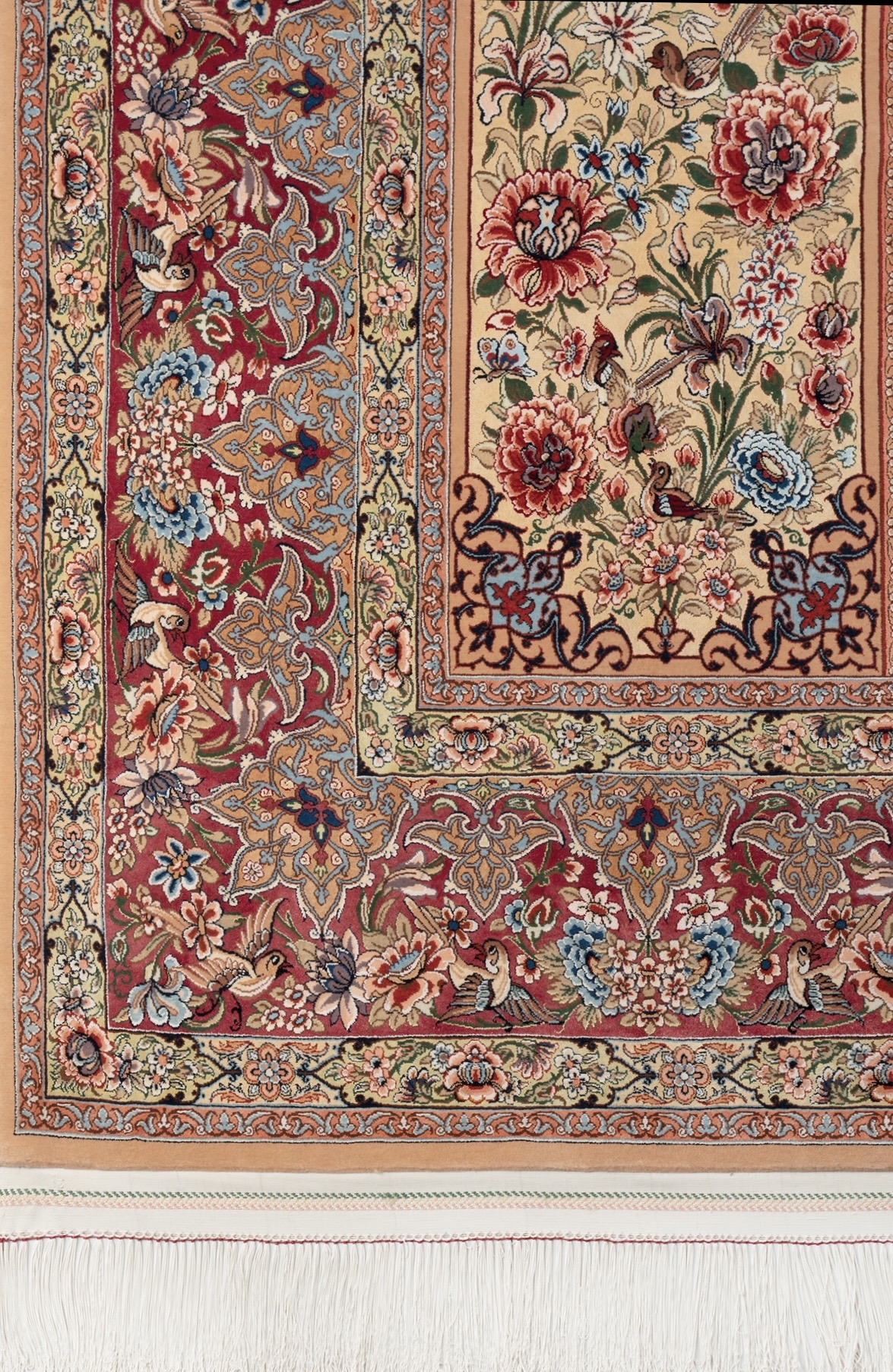 A corner of the piece, the crimson border of palmette vinework and birds turning at the corner, the cream field beyond carrying peony and iris among small birds, with the silk fringe below.