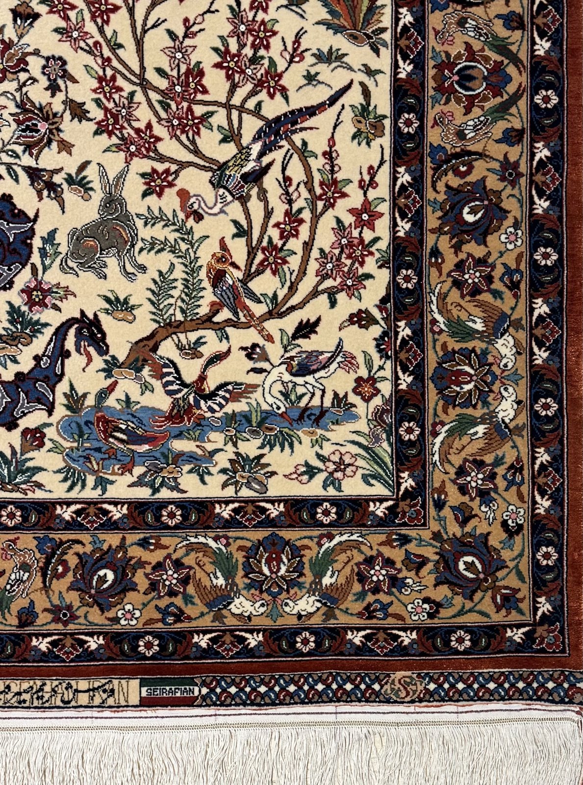 The lower corner of the carpet, with the woven Seirafian signature in its cartouche, the layered borders, a hare and a heron at the water.