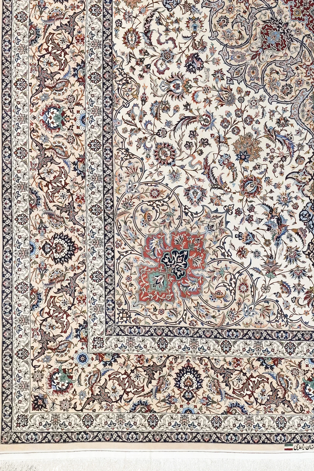 The corner of one carpet of the pair: the camel main border and navy guards turning the angle, a rose corner medallion on the ivory field, and the woven signature Iran, Isfahan, Zojaji between two flags above the silk fringe.